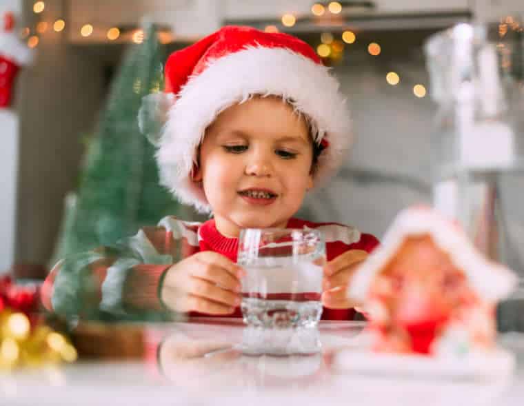 Happy funny toddler boy in a red Santa hat drinking filtered water from a glass in the kitchen. Holidays, health concept.