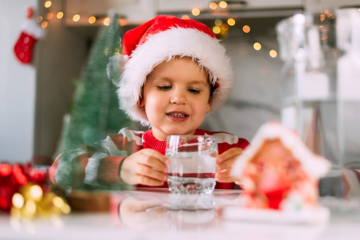 Happy funny toddler boy in a red Santa hat drinking filtered water from a glass in the kitchen. Holidays, health concept.