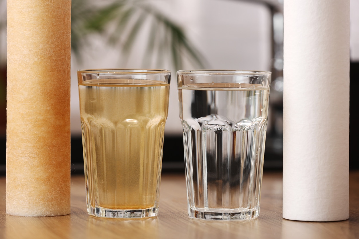 House water filtration system to drinkable condition. New clean water filter cartridge and glass of clear water. Used cartridge in rust and glass of dirty water on tabletop kitchen.