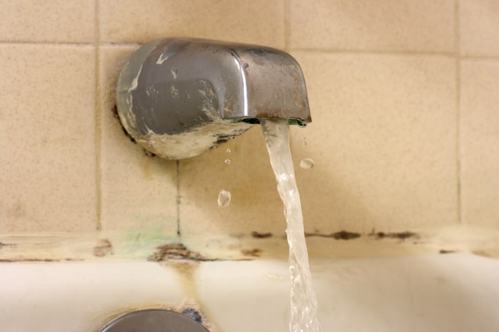 Selective focus on old bath tub faucet with water flowing close up showing rusty metal, toxic black mold (Stachybotrys atra) on grout, and need for new bathroom renovation